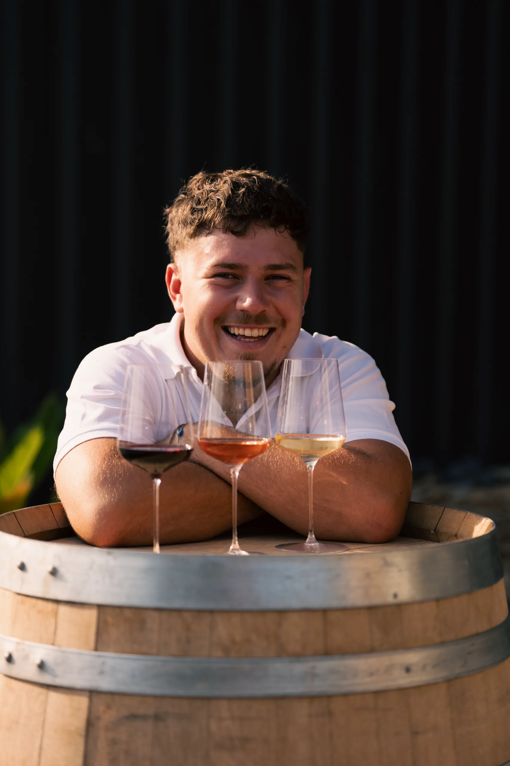 Numa Rouvier souriant, accoudé à un fût avec trois verres de vin